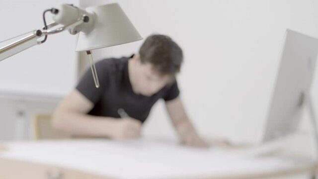 Young man in a black t-shirt is seen sitting at a desk sketching / drawing.