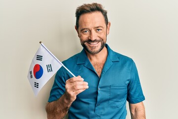 Middle age man holding south korea flag looking positive and happy standing and smiling with a confident smile showing teeth