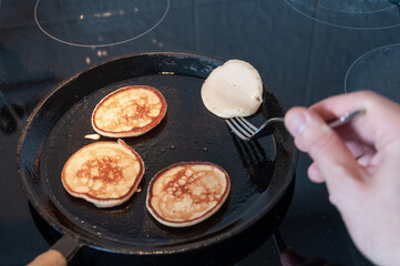 A man makes pancakes in a cast-iron skillet. In his hand he holds a fork and uses it to turn the pancake.