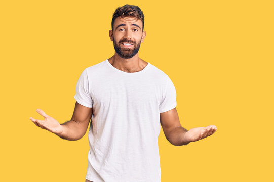 Young hispanic man wearing casual white tshirt clueless and confused expression with arms and hands raised. doubt concept.
