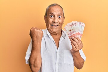 Handsome mature man holding 10 colombian pesos banknotes screaming proud, celebrating victory and...