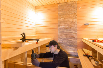 Sauna construction, finishing. The man is screwing a wooden bench to the wall. - obrazy, fototapety, plakaty