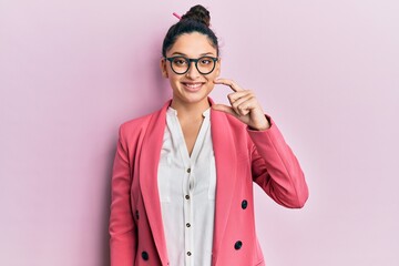 Beautiful middle eastern woman wearing business jacket and glasses smiling and confident gesturing with hand doing small size sign with fingers looking and the camera. measure concept.