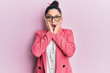 Beautiful middle eastern woman wearing business jacket and glasses afraid and shocked, surprise and amazed expression with hands on face