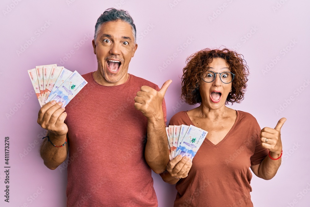 Poster beautiful middle age couple holding colombian pesos banknotes pointing thumb up to the side smiling 