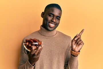 Young african american man holding bowl with dates smiling happy pointing with hand and finger to the side