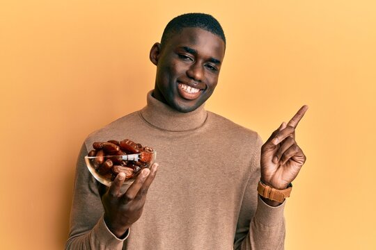 Young african american man holding bowl with dates smiling happy pointing with hand and finger to the side