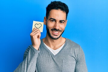 Young hispanic man holding heart reminder looking positive and happy standing and smiling with a confident smile showing teeth