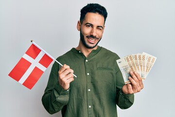 Young hispanic man holding denmark flag and krone banknotes winking looking at the camera with sexy...