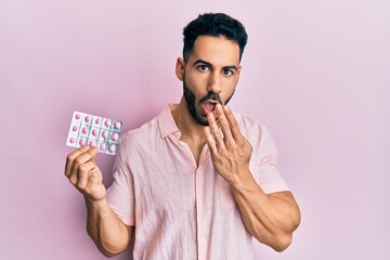 Young hispanic man holding pills covering mouth with hand, shocked and afraid for mistake....