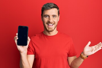 Handsome caucasian man holding smartphone showing screen celebrating achievement with happy smile and winner expression with raised hand