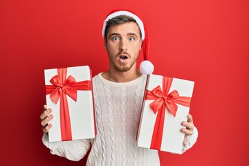 Handsome caucasian man wearing christmas hat and holding gifts afraid and shocked with surprise and...