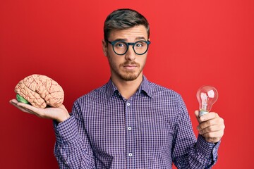 Handsome caucasian man holding brain and lightbulb for inspiration and idea skeptic and nervous,...