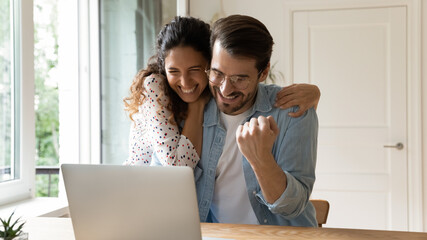 Laughing excited millennial family couple embracing by computer screen winning prize at lottery or...