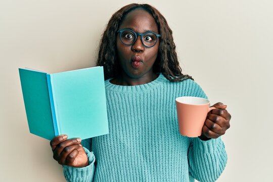 African young woman reading a book and drinking a cup of coffee making fish face with mouth and squinting eyes, crazy and comical.