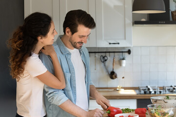 Grateful young wife embracing shoulder of husband engaged in preparing tasty healthy vegetable salad. Young man boyfriend skilled in cooking food doing delicious meal to treat beloved woman girlfriend