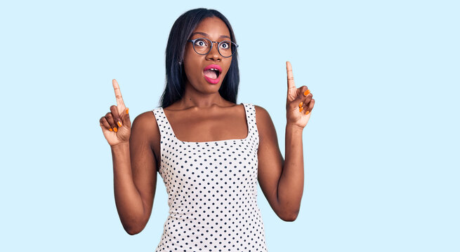 Young african american woman wearing casual clothes and glasses amazed and surprised looking up and pointing with fingers and raised arms.