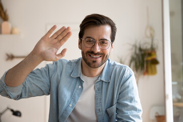 Smiling millennial male in glasses posing before digital webcam waving hand chatting with friend...