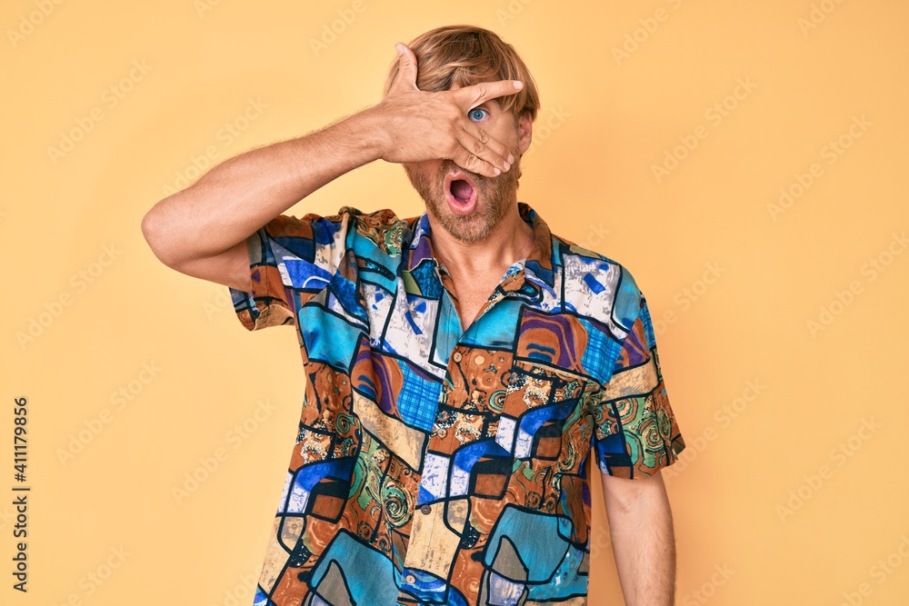 Poster young blond man wearing summer shirt peeking in shock covering face and eyes with hand, looking thro