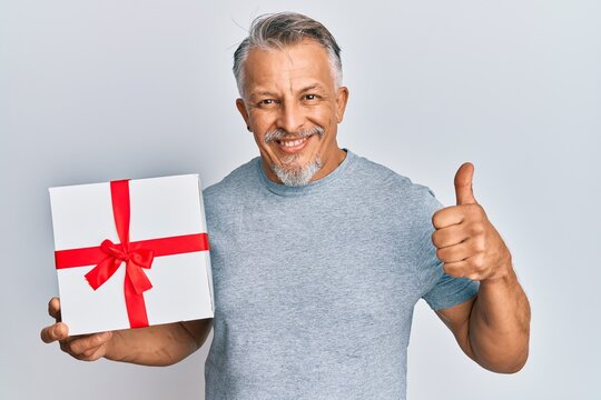 Middle age grey-haired man holding gift smiling happy and positive, thumb up doing excellent and approval sign