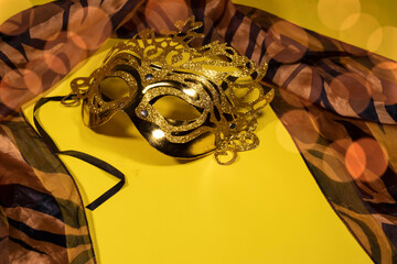 On a yellow background with bokeh, there is a silk scarf and a gilded carnival mask.
