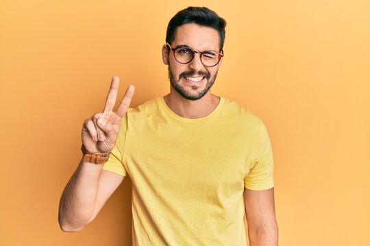 Young hispanic man wearing casual clothes and glasses smiling with happy face winking at the camera doing victory sign. number two.
