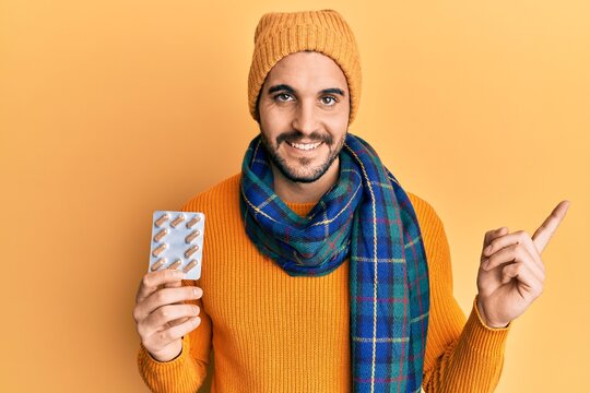 Young hispanic man wearing wool sweater and winter scarf holding pills smiling happy pointing with hand and finger to the side