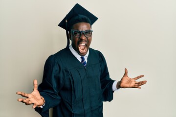 Handsome black man wearing graduation cap and ceremony robe crazy and mad shouting and yelling with...