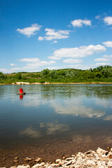 View from the river bank to the red buoy on a bright summer day. The red buoy is reflected in the calm water of the river. Blue sky and clouds are reflected in the river.