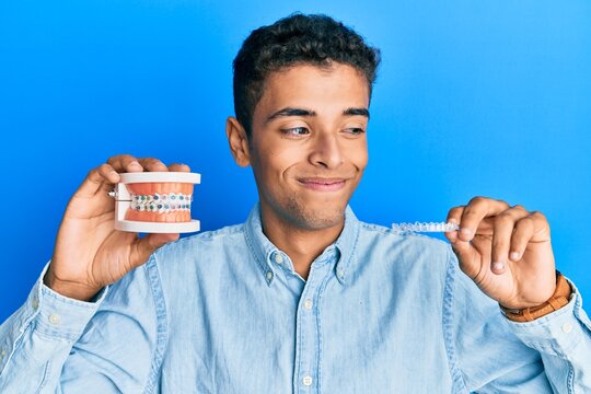 Young handsome african american man holding invisible aligner orthodontic and braces smiling looking to the side and staring away thinking.