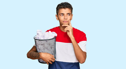 Young handsome african american man holding paper bin full of crumpled papers serious face thinking...