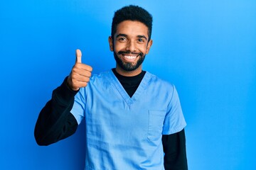 Fototapeta premium Handsome hispanic man with beard wearing blue male nurse uniform smiling happy and positive, thumb up doing excellent and approval sign