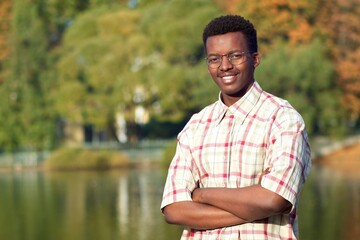 Portrait of handsome happy black African Afro American young man standing in shirt in a park near lake, smiling with his hands crossed. Ethiic student.