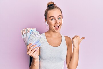 Beautiful blonde sport woman holding 20 swedish krona banknotes pointing thumb up to the side smiling happy with open mouth