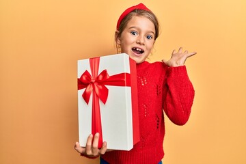 Little beautiful girl holding gift celebrating achievement with happy smile and winner expression with raised hand