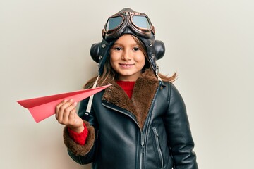 Little beautiful girl wearing pilot uniform holding paper plane looking positive and happy standing and smiling with a confident smile showing teeth