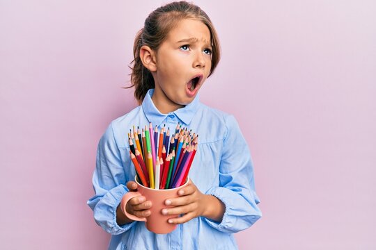 Little beautiful girl holding colored pencils angry and mad screaming frustrated and furious, shouting with anger. rage and aggressive concept.