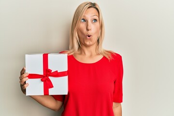 Beautiful middle age blonde woman holding gift scared and amazed with open mouth for surprise, disbelief face