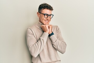 Hispanic young man wearing casual turtleneck sweater laughing nervous and excited with hands on chin looking to the side