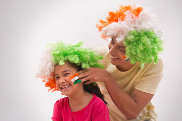 young man painting flag on a girls cheek wearing tricolour wig, independence day	
