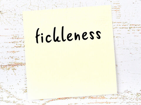 Yellow sticky note on wooden wall with handwritten word fickleness