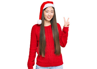 Young chinese woman wearing christmas hat smiling looking to the camera showing fingers doing...