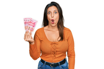 Beautiful hispanic woman holding 100 yuan chinese banknotes scared and amazed with open mouth for surprise, disbelief face