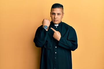 Young latin priest man standing over yellow background in hurry pointing to watch time, impatience,...