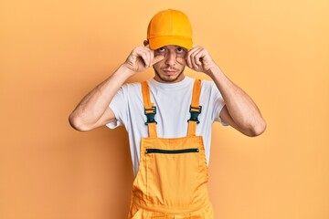 Hispanic young man wearing handyman uniform trying to open eyes with fingers, sleepy and tired for morning fatigue