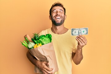 Handsome man with beard holding groceries and 1 american dollar banknote smiling and laughing hard...