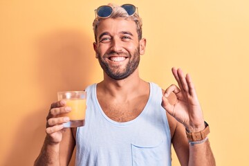 Young handsome blond man with beard on vacation drinking glass of healthy orange juice doing ok...