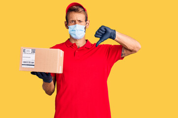 Fototapeta na wymiar Handsome blond man with beard holding delivery box wearing medical mask with angry face, negative sign showing dislike with thumbs down, rejection concept