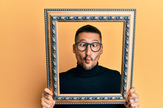 Handsome man with tattoos holding empty frame making fish face with mouth and squinting eyes, crazy and comical.