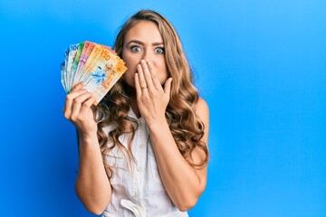 Young blonde girl holding swiss franc banknotes covering mouth with hand, shocked and afraid for mistake. surprised expression
